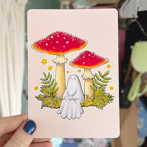 May include: A white ghost with a friendly face sits between two red and white mushrooms with white spots. The mushrooms are surrounded by green ferns and yellow stars.