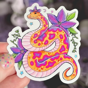 May include: A sticker featuring a cartoon snake wearing a witch's hat. The snake is pink, orange, and purple with a spotted pattern. The snake is curled up with a purple flower behind it. There are stars and a crescent moon in the background.