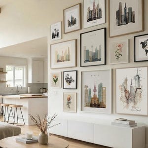 The image is a modern living room with a gallery wall featuring various framed artworks depicting cityscapes and landscapes. The room has a clean, minimalist aesthetic with white walls, a wooden floor, and a white sofa.