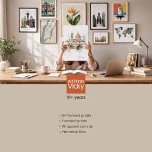 A person is sitting at a desk in a room, holding up a framed print of a cityscape. The desk is covered with various items, including books, a laptop, and a potted plant. The wall behind the desk is decorated with multiple framed prints, including a map and a city skyline. The image also includes text that describes the products offered by ArtPrints Vicky, such as "Unframed prints", " Framed prints", " Wrapped canvas", and "Printable files".