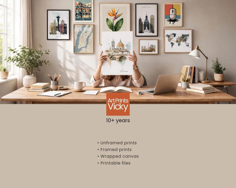 A person is sitting at a desk in a room, holding up a framed print of a cityscape. The desk is covered with various items, including books, a laptop, and a potted plant. The wall behind the desk is decorated with multiple framed prints, including a map and a city skyline. The image also includes text that describes the products offered by ArtPrints Vicky, such as "Unframed prints", " Framed prints", " Wrapped canvas", and "Printable files".