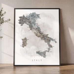May include: Framed map of Italy with a watercolor effect in shades of gray, beige, and white. The map is detailed with city names and is labeled "ITALY" at the bottom. The frame is black.