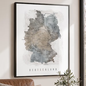 May include: A framed wall art print featuring a watercolor map of Germany in shades of gray, blue, and brown. The map displays state names and major cities with black text labels. The word DEUTSCHLAND is printed in black at the bottom center.