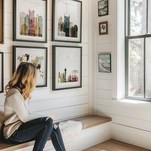 A woman sits on a bench in a room with framed cityscape art on the walls, looking out a window.
