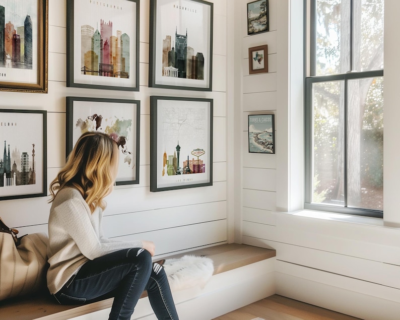 A woman sits on a bench in a room with framed cityscape art on the walls, looking out a window.