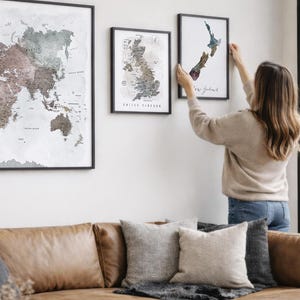 A woman hangs two framed world maps on a wall in a living room.
