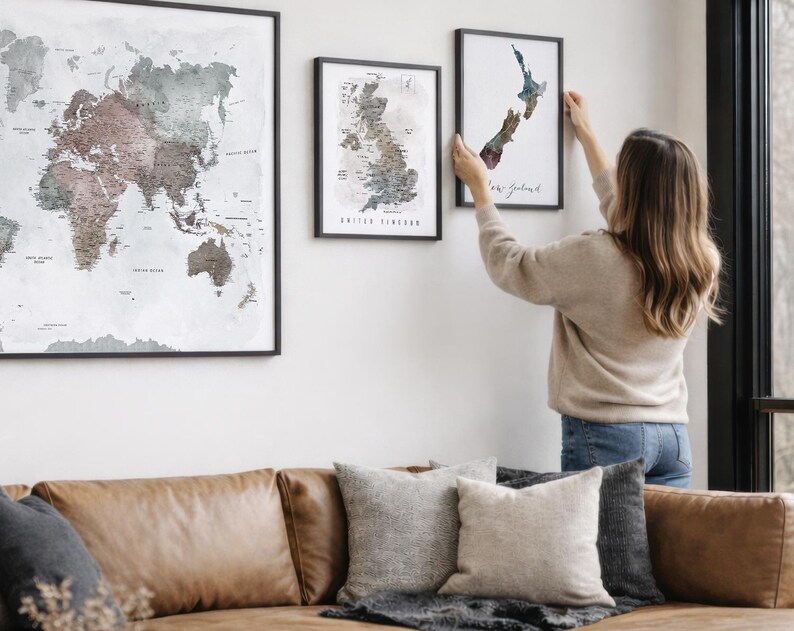 A woman hangs two framed world maps on a wall in a living room.