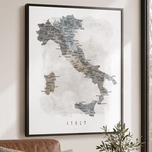 May include: A framed wall art print featuring a watercolor-style map of Italy in shades of gray and muted brown. The map displays regional names and city locations with black text. The word ITALY is printed in a simple sans-serif font at the bottom.