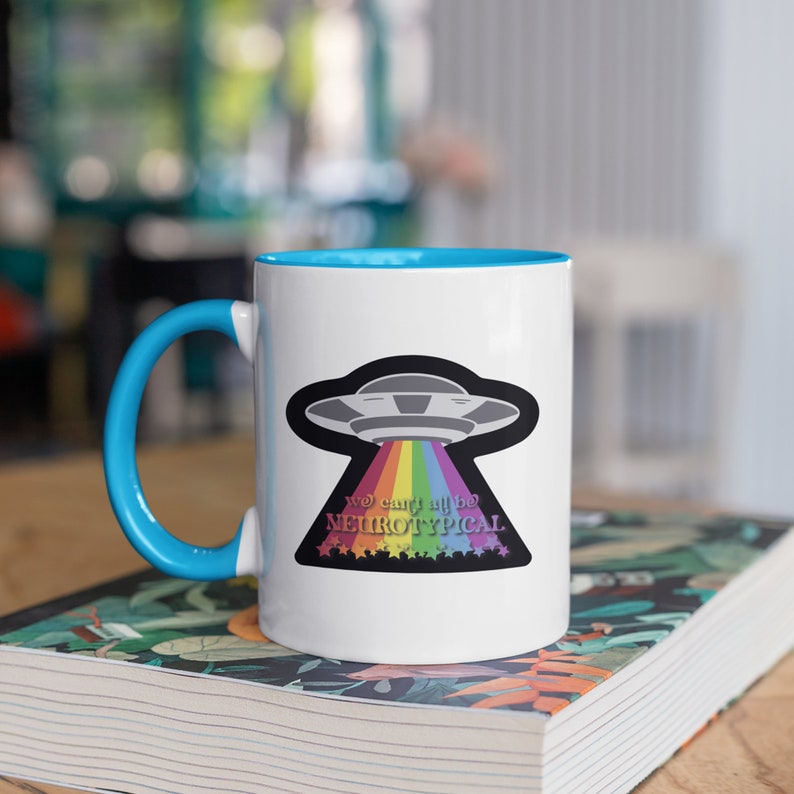 May include: White ceramic mug with a blue rim and handle. The mug features a black and white illustration of a UFO with a rainbow beam shooting out of it. The text "You can't be neurotypical" is written below the UFO.