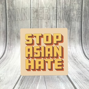 May include: Square sticker with the text "STOP ASIAN HATE" in yellow and orange, with a shadow effect. The sticker has a light tan background. The sticker is on a light grey wooden surface.