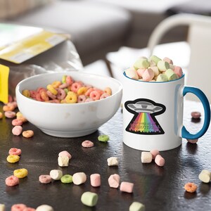 May include: White ceramic mug with a blue rim and handle. The mug has a graphic of a UFO with a rainbow coming out of it. The text on the mug reads "I'm sure all of us are neurotypical".