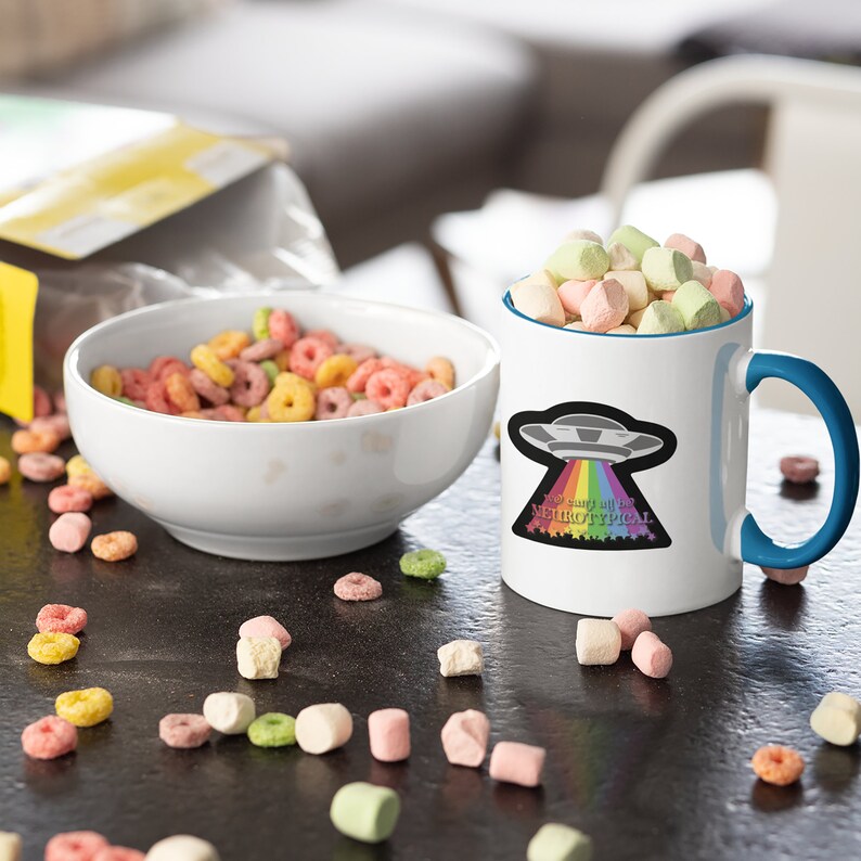 May include: White ceramic mug with a blue rim and handle. The mug has a graphic of a UFO with a rainbow coming out of it. The text on the mug reads "I'm sure all of us are neurotypical".