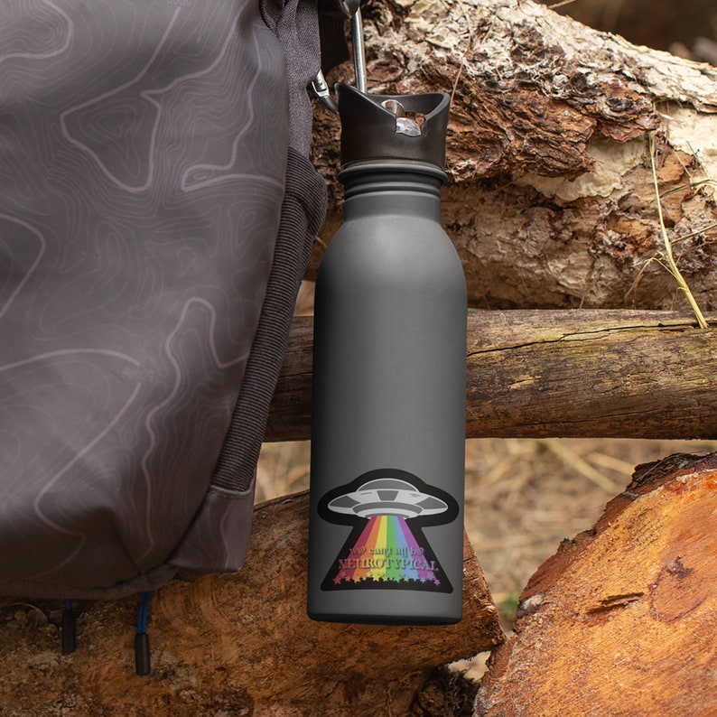 May include: A gray reusable water bottle with a black lid and a rainbow sticker that says "So smart, it's be neurotypical". The sticker features a cartoon UFO with a rainbow beam of light coming out of it.