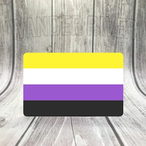 May include: A rectangular sticker featuring the non-binary pride flag. The flag design includes horizontal stripes in yellow, white, purple, and black. The sticker has rounded corners and is set against a wooden background.