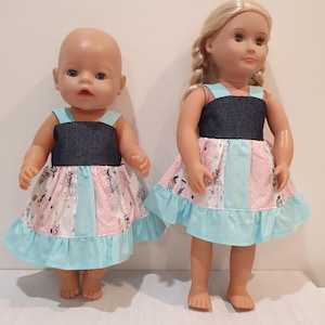 May include: Two dolls dressed in matching outfits. The dresses feature a dark blue bodice and a skirt with a pink and white pattern of bears and feathers. A light blue ruffle trims the hem of the skirt.