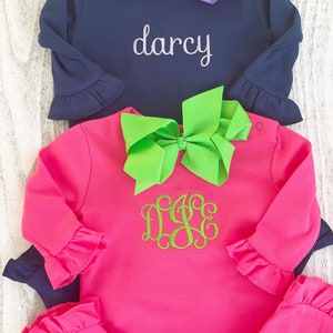 May include: Two long-sleeved shirts with ruffled sleeves. The top shirt is navy blue with the name "darcy" embroidered on the front. The bottom shirt is pink with a green monogram on the front. Both shirts have a bow on the front.