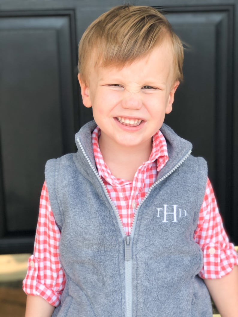 May include: A young boy wearing a grey fleece vest with a white monogram "THD" on the front. He is also wearing a red and white gingham button-down shirt.