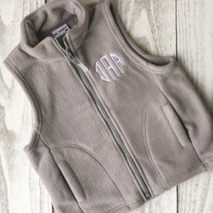 May include: A grey fleece vest with a zip fastening and a white monogram on the front. The vest has a pocket on the right side.