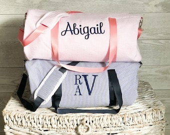 May include: Two striped canvas duffel bags with pink and black straps. The top bag is pink with white stripes and is embroidered with the name "Abigail". The bottom bag is blue with white stripes and is embroidered with the initials "R.A.V."