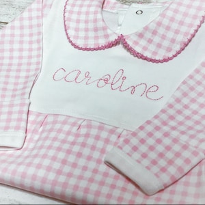 May include: Pink and white gingham baby onesie with a white collar and the name "caroline" embroidered on the front.
