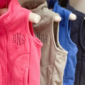 May include: Four fleece vests in different colours, hanging on hangers. The vests have zipper closures and are embroidered with the letters "ENG", "MILA", and two other letters.