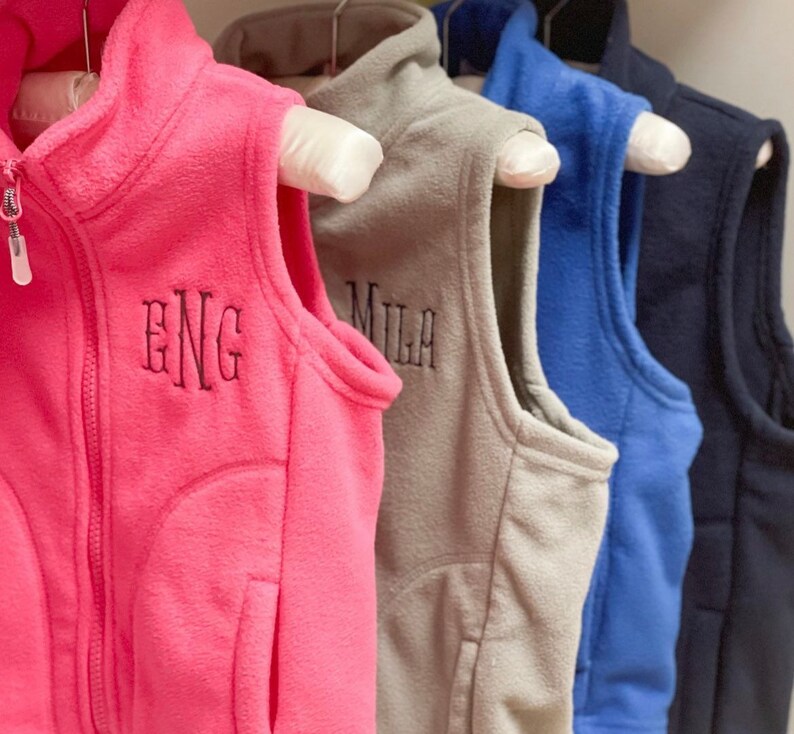 May include: Four fleece vests in different colours, hanging on hangers. The vests have zipper closures and are embroidered with the letters "ENG", "MILA", and two other letters.