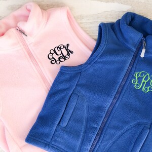 May include: Two fleece vests, one pink with black embroidery and the other blue with green embroidery. Both vests have a zip closure and a stand-up collar.