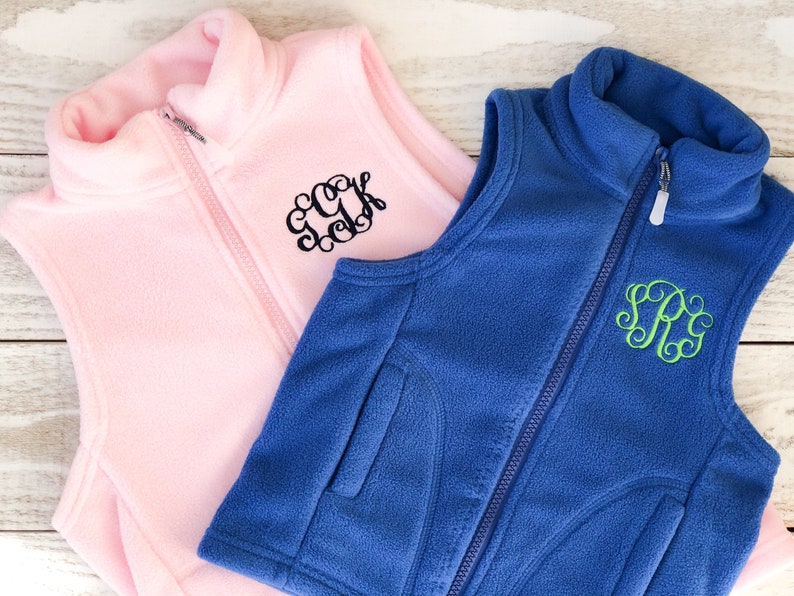 May include: Two fleece vests, one pink with black embroidery and the other blue with green embroidery. Both vests have a zip closure and a stand-up collar.