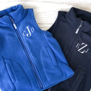 May include: Two fleece vests with zip fastenings. The vest on the left is blue with white embroidered initials "CJD". The vest on the right is navy blue with white embroidered initials "EZJ".