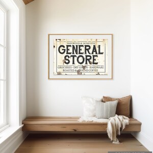 May include: A vintage-style poster print with a rustic, weathered background featuring the text "IMPORTED & DOMESTIC GENERAL STORE GROCERIES DRY GOODS HARDWARE ROASTED & GROUND COFFEE". The poster is framed in a simple, wooden frame and is displayed on a wall above a wooden bench.