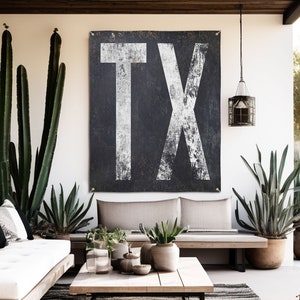 May include: A black and white canvas wall art featuring the letters "TX" in a distressed, vintage style. The canvas is hanging on a white wall in a living room setting with plants and a couch.