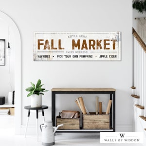 May include: A white sign with brown text that reads "Little Farm Fall Market Every Weekend Hayrides Pick Your Own Pumpkins Apple Cider". The sign is hanging on a white wall above a black table with a wooden crate on it.