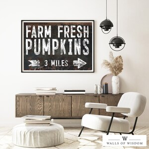 May include: A black and white sign with the text "Farm Fresh Pumpkins" and an arrow pointing to the right. The sign also says "3 Miles". The sign is hanging on a white wall in a living room setting.