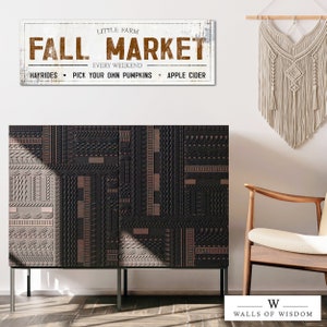 May include: A white sign with brown text that reads "Little Farm Fall Market Every Weekend Hayrides Pick Your Own Pumpkins Apple Cider". The sign is hanging on a white wall above a black cabinet with a geometric pattern. A wooden chair with a white cushion is in the foreground.