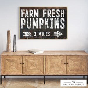 May include: A rustic black and white sign with the text "FARM FRESH PUMPKINS 3 MILES" and a white arrow pointing to the right. The sign is framed in a brown wood frame and is hanging on a white wall above a wooden cabinet.