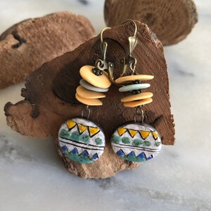 Pu&ograve; includere: Un paio di orecchini pendenti con un motivo geometrico in tonalit&agrave; di blu, verde, giallo e bianco. Gli orecchini sono in ceramica e hanno un aspetto rustico e fatto a mano.
