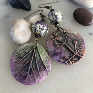 Pode incluir: Brincos artesanais com um design de folha em tons de roxo e verde. Os brincos têm uma aparência texturizada e são acentuados com pequenas contas estampadas. Os componentes metálicos têm um acabamento em latão antigo.
