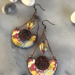 May include: A pair of dangle earrings with a floral pattern and a brown metal circle. The earrings have a chain with a beige bead and a red bead.