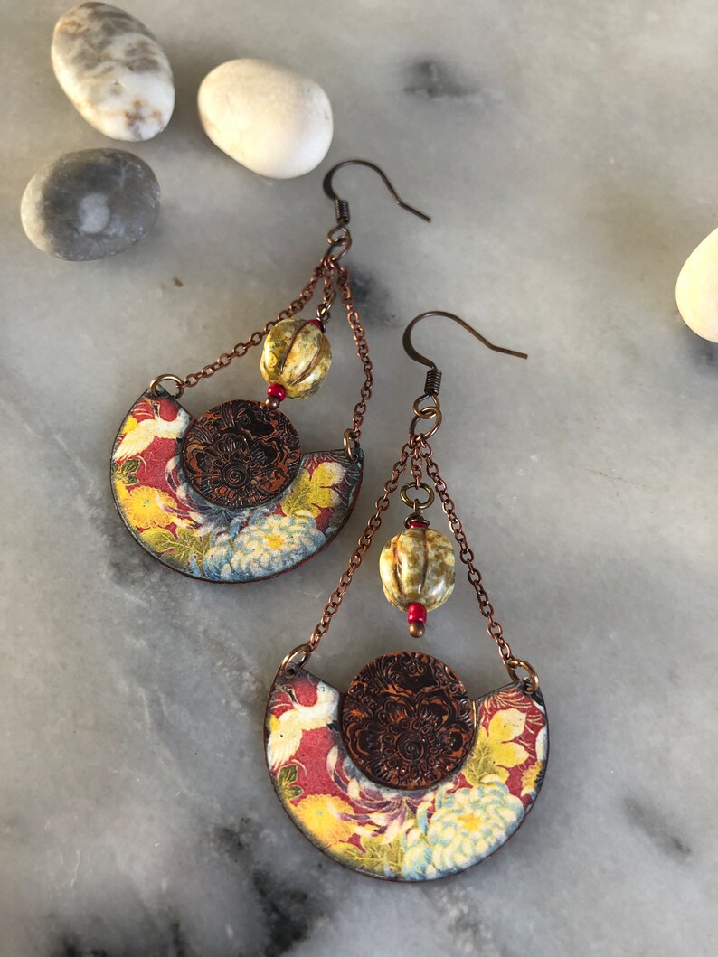 May include: A pair of dangle earrings with a floral pattern and a brown metal circle. The earrings have a chain with a beige bead and a red bead.