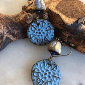 Op de afbeelding: Een paar blauwe en gouden keramische oorbellen met een gedetailleerd bloemenmotief. De oorbellen hangen aan een gouden haak en hebben een kleine blauwe kraal.