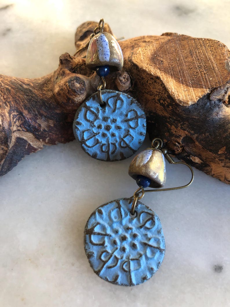 Op de afbeelding: Een paar blauwe en gouden keramische oorbellen met een gedetailleerd bloemenmotief. De oorbellen hangen aan een gouden haak en hebben een kleine blauwe kraal.