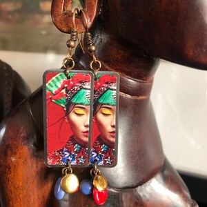 May include: Copper-toned dangle earrings. Each earring features a rectangular pendant with a woman's portrait in traditional attire against a red background. The earrings have blue, red, and gold-colored beads.