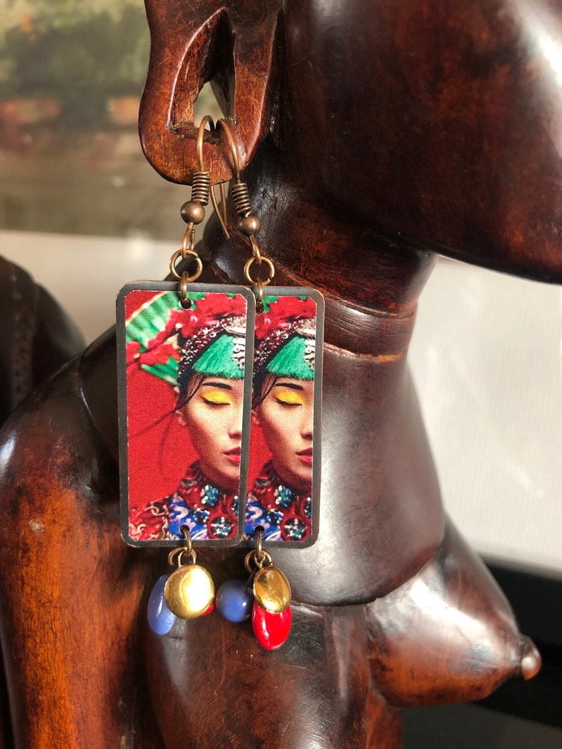 May include: Copper-toned dangle earrings. Each earring features a rectangular pendant with a woman's portrait in traditional attire against a red background. The earrings have blue, red, and gold-colored beads.