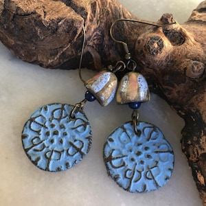 Op de afbeelding: Een paar blauwe en gouden oorbellen met een bloemmotief. De oorbellen zijn gemaakt van keramiek en hebben een verweerde afwerking. De oorbellen hangen aan een bruin houten oppervlak.