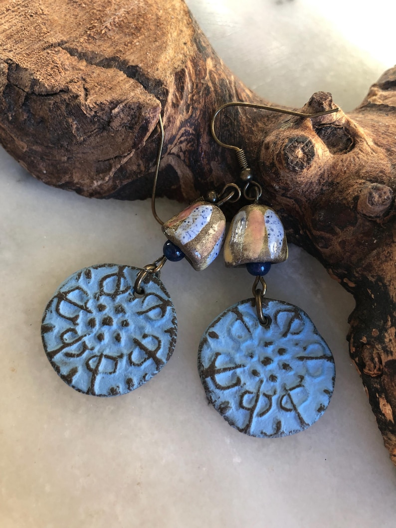 Op de afbeelding: Een paar blauwe en gouden oorbellen met een bloemmotief. De oorbellen zijn gemaakt van keramiek en hebben een verweerde afwerking. De oorbellen hangen aan een bruin houten oppervlak.