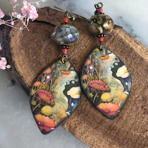 Pode incluir: Um par de brincos pendentes com pingentes de metal em forma de folha com uma estampa floral colorida em tons de laranja, amarelo e verde. Cada brinco é acentuado com uma conta de vidro redonda multicolorida e pequenas contas vermelhas.