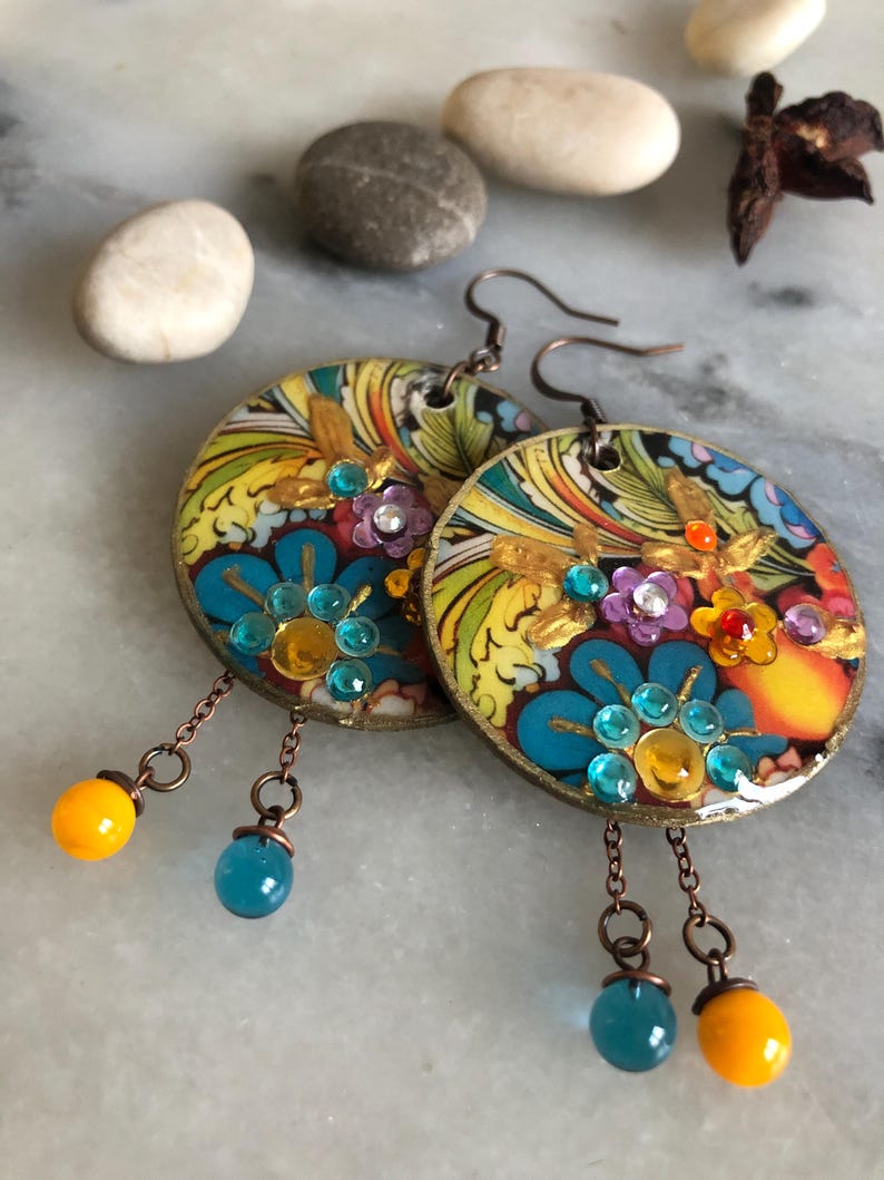 K&ouml;nnte beinhalten: Ein Paar handgefertigte, runde Ohrringe mit einem farbenfrohen Blumenmuster. Die Ohrringe haben blaue, gelbe und lila Edelsteine mit h&auml;ngenden orangefarbenen und blauen Perlen. Hergestellt aus kupferfarbenem Metall.