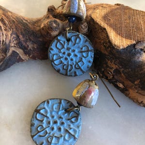 Op de afbeelding: Een paar hangende oorbellen met blauwe en bruine keramische accenten. De oorbellen hebben een ronde, getextureerde blauwe keramische schijf met een kleinere, ronde, bruine keramische schijf met witte en roze accenten die eronder hangen.