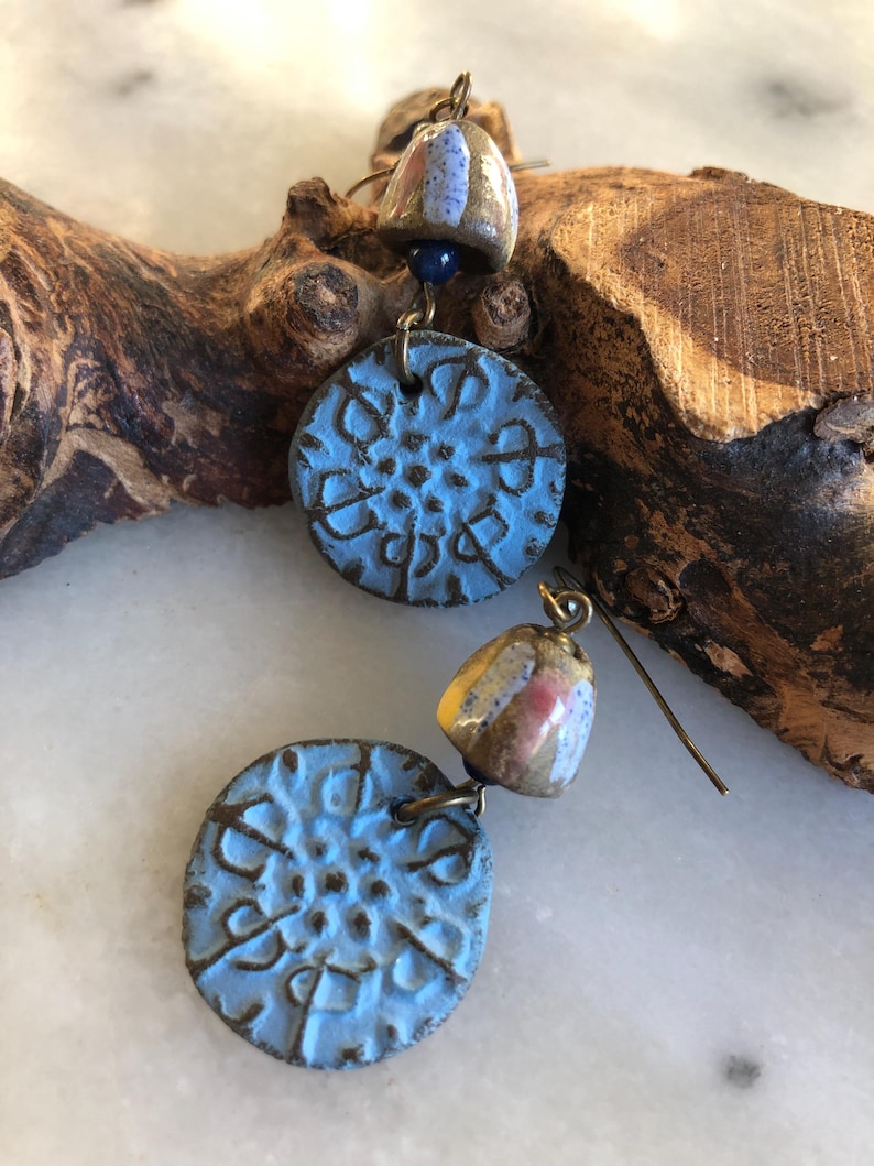 Op de afbeelding: Een paar hangende oorbellen met blauwe en bruine keramische accenten. De oorbellen hebben een ronde, getextureerde blauwe keramische schijf met een kleinere, ronde, bruine keramische schijf met witte en roze accenten die eronder hangen.