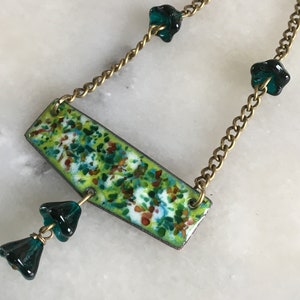 Op de afbeelding: Een vintage ketting met een rechthoekige hanger met een gespikkeld groen, wit en oranje emaille ontwerp. De goudkleurige ketting heeft donkergroene bloemvormige kralen. De ketting is een uniek sieraad.
